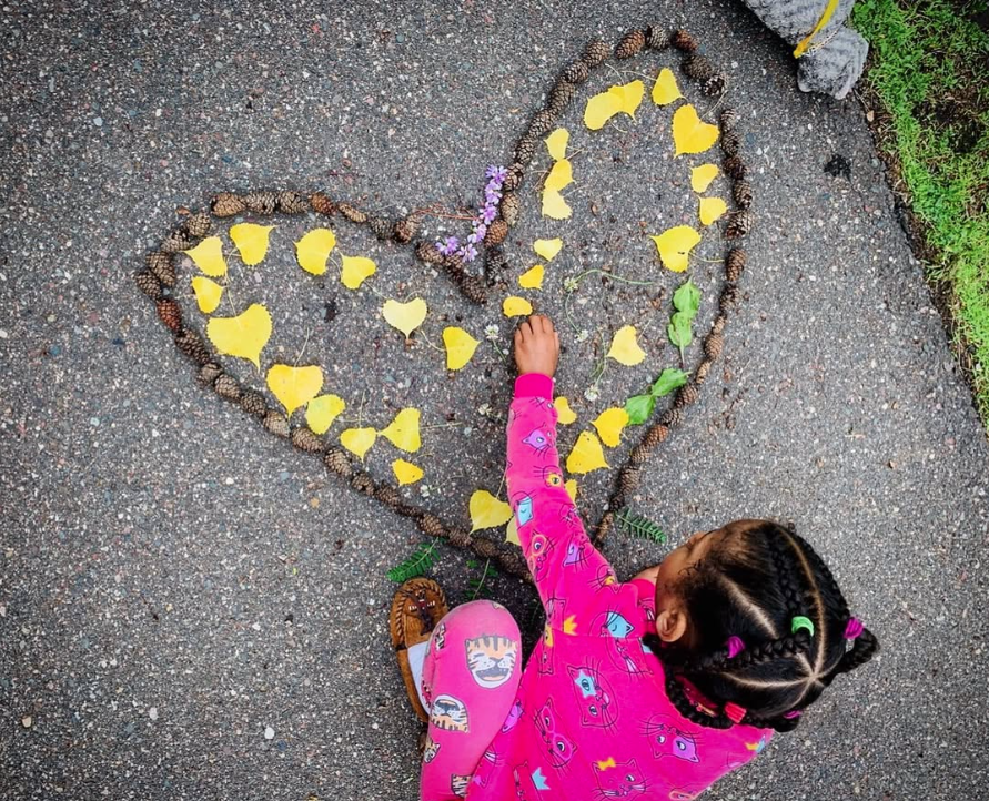 child creating heart on sidewalk with pinecones and leaves